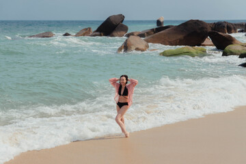 A woman in a swimsuit and light shirt stands on a sandy beach, smiling and enjoying the warm sun and waves. The beach is backed by large rocks and turquoise waters