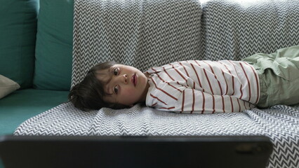 Young boy lying on a patterned couch, watching a tablet screen with intent, relaxed and engaged in a home setting, showcasing the use of technology for entertainment and learning