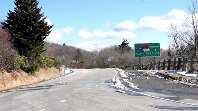 Roadside view featuring a Welcome to North Carolina sign amidst a winter landscape with snow patches and clear blue sky. High quality 4k footage