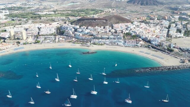 Panorama flight over sailing yachts, blue Atlantic Ocean, beach Playa de Las Vistas and hotels towards volcano Montana Chayofita in holiday resort Los Cristianos on Canary Island Tenerife, Spain