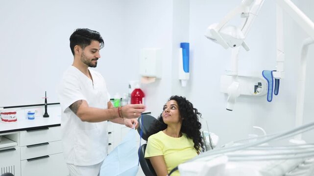 Dentist putting dental bib around patients neck. Slow motion of young Hispanic male stomatologist putting patient bib around neck of woman with curly hair sitting on dental chair