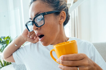 One adult woman yawn at home in daylight sitting on sofa holding a cup of coffee to wake up from...