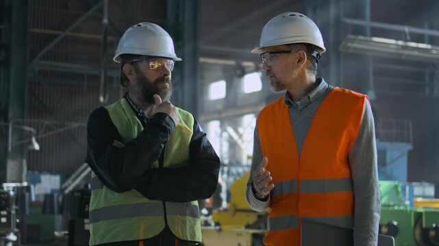 Two professional heavy industry engineers in safety uniform and hard hats discuss work process at manufacturing factory. Mature supervisor talks to adult worker at plant or workshop. Slow motion.