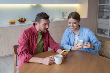 Interested family couple looking at screen cellphone have fun together in morning. Joyful happy woman and pleased man spend free time weekend enjoy coffee drink, surf internet, scroll social networks.