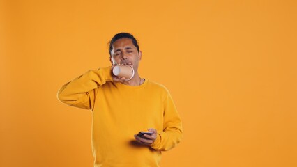 Indian man enjoying fresh coffee from disposable paper cup early in the morning to wake up and be energized. Person drinking hot beverage from recycled takeaway cup, studio background, camera B