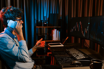 Working young business trader discussing with broker on smartphone with market stock graph in real time monitor screens at neon light modern office. Concept of analyzing dynamic exchange rate. Gusher.
