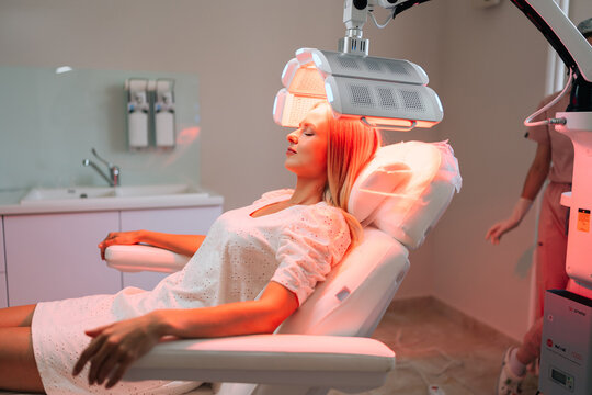 Side view of woman client with blonde hair lying chair having LED light hair PDT treatment at luxury aesthetic clinic. Female specialist adjusting innovation device for photodynamic therapy procedure