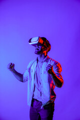 Professional gamer put the hand in the air to celebrate for winning game while wearing VR goggle at colorful neon light background. Caucasian man using technology innovation and enter. Deviation.