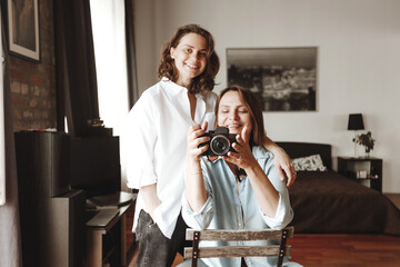 Two young smiling women with a camera in a living room. They are happy and looking at photos or taking pictures.