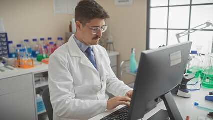 A young hispanic man in a lab coat works attentively on a computer in a laboratory setting.