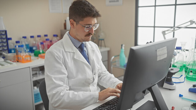A young hispanic man in a lab coat works attentively on a computer in a laboratory setting.