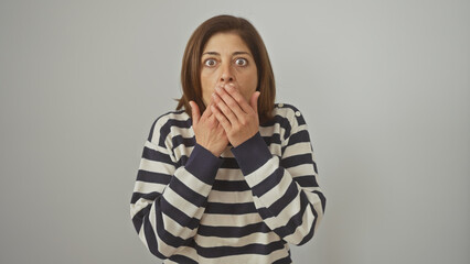 Shocked middle-aged woman covers mouth with hands against a white background, wearing a striped...