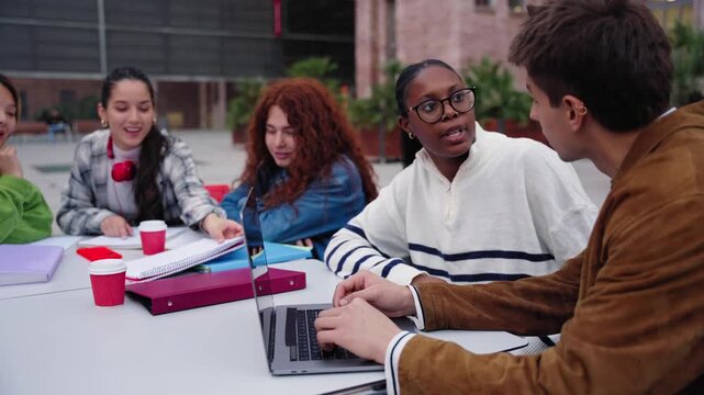 Cheerful group of diverse generation z students working together sitting at table on university campus terrace. Multiracial young classmates gathered together using laptop outdoors