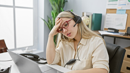 Stressed young woman with blonde hair wearing headset in modern office setting, expressing concern.
