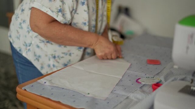 A senior woman traces a paper pattern onto striped fabric using chalk. working on a table with various sewing tools, including a measuring tape and pins