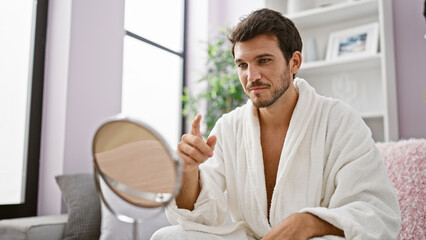 A young bearded man in a white bathrobe engages with his reflection in a cozy home setting.