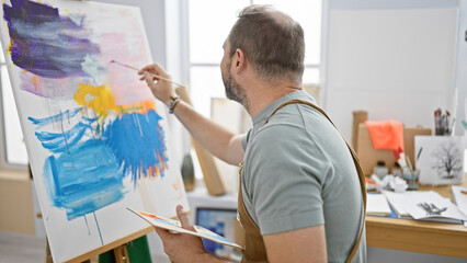 Mature hispanic man with grey hair painting on canvas in a bright art studio, conveying creativity and artistry.