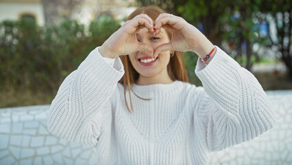 A cheerful young woman forms a heart with her hands in a sunny park setting, symbolizing love and happiness.