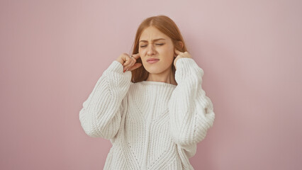 Young redhead woman covering ears isolated over pink background, showing discomfort or noise...