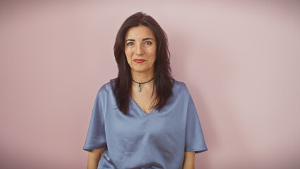 A confident middle-aged hispanic woman stands isolated against a pink wall background, portraying a mature and beautiful presence.