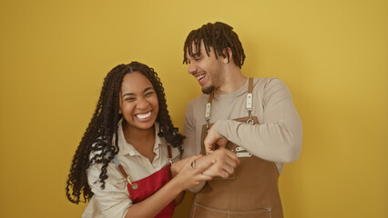 A man and woman in aprons laugh together against a yellow backdrop, suggesting a cheerful work environment.