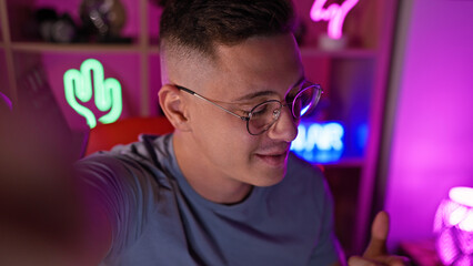 Smiling young hispanic man wearing glasses in a colorful neon-lit gaming room at night.
