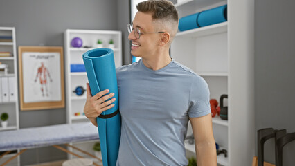 Smiling hispanic man holding yoga mat in a therapy clinic, portraying health and wellness.
