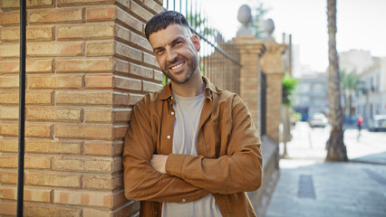Fototapeta premium Smiling young hispanic man with beard, standing cross-armed on an urban street, wearing casual clothes.