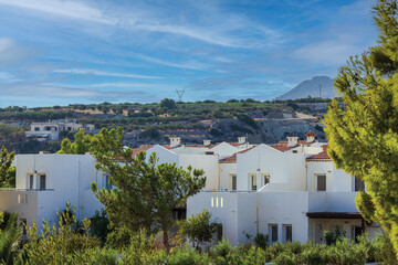 Fototapeta premium Beautiful view of white two-story hotel buildings with tropical garden plants and mountain pines, set against mountain backdrop on Crete.