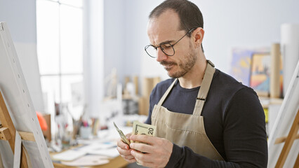 A bearded bald man in glasses examines money indoors, wearing a black shirt and apron against an...
