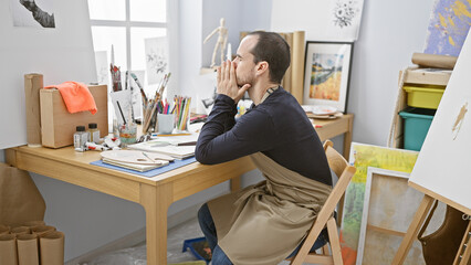 Pensive bearded man in apron sitting at a desk filled with artistic supplies in a well-lit studio