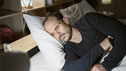 Fototapeta premium Hispanic bald man with beard sleeps uncomfortably in a dark bedroom, showing insomnia.