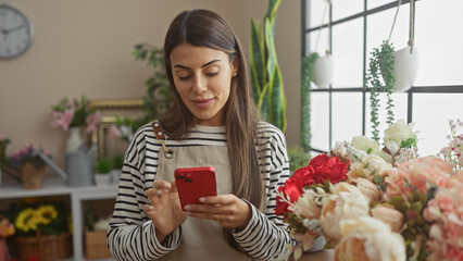 Hispanic woman using smartphone indoors surrounded by flowers in a well-lit room.