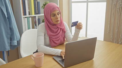 A young woman wearing a hijab examines her credit card while using a laptop in a well-lit home interior.