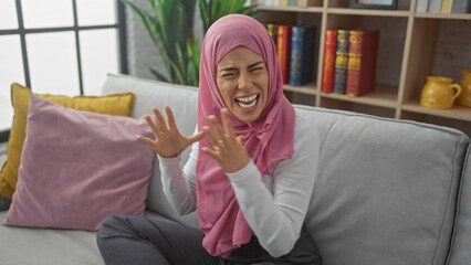 Stressed young woman shouting, in hijab indoors, showing frustration in a living room setting.