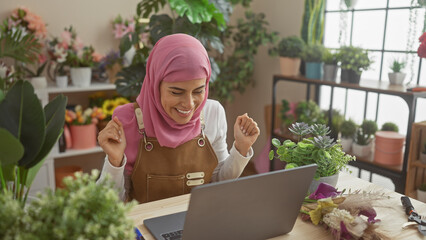 A smiling woman wearing a hijab arranges flowers joyfully at home, using a laptop in a cozy floral shop setting.