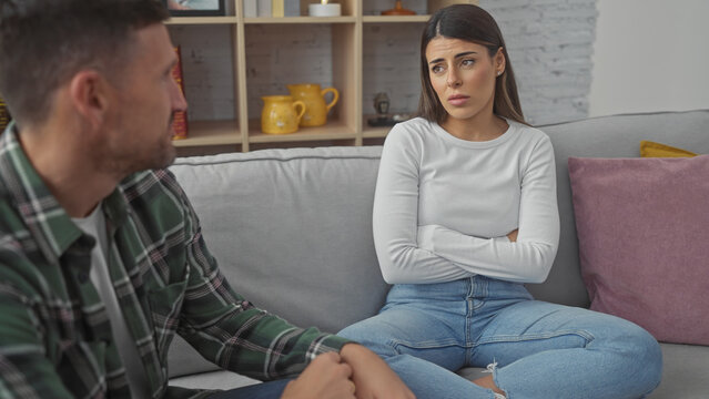 An upset hispanic woman and a comforting man sitting together on a sofa in a cozy living room, implying relationship issues.