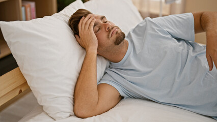 A young hispanic man with a beard appears in pain, lying in bed indoors, clutching his head.