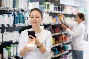 Positive woman customer using phone to search information about selected product at pharmacy