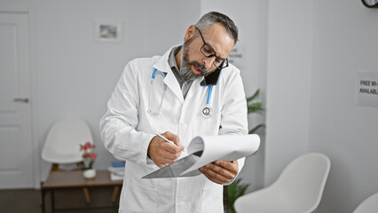 Mature, bearded, hispanic doctor multitasks in clinic lobby with phone and paperwork.