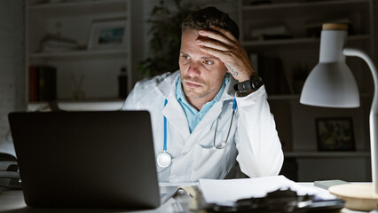 Tired hispanic male doctor in lab coat working late in the hospital office with laptop and paperwork