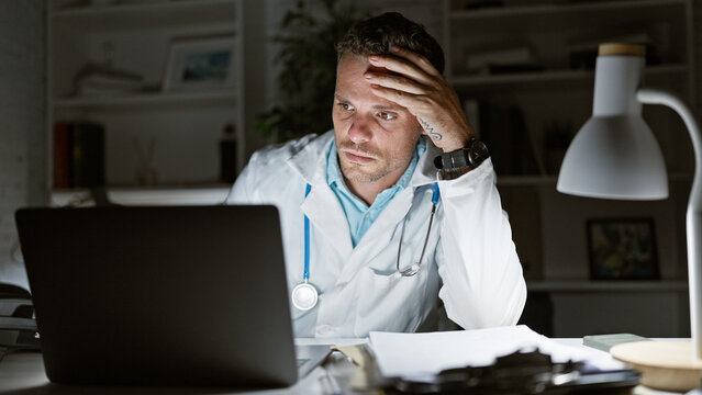 Tired hispanic male doctor in lab coat working late in the hospital office with laptop and paperwork