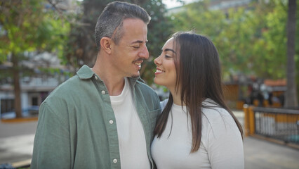 Naklejka premium A smiling man and woman lovingly gaze at each other, outdoors in a sunny, tree-lined urban park.