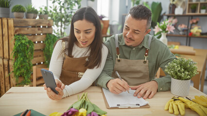 A woman and man, team of florists, are in a flower shop working together, with the woman holding a smartphone and the man writing on a clipboard.