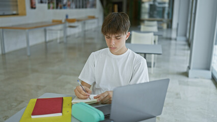 Young caucasian male student focused on writing in notebook at table with laptop in university library.