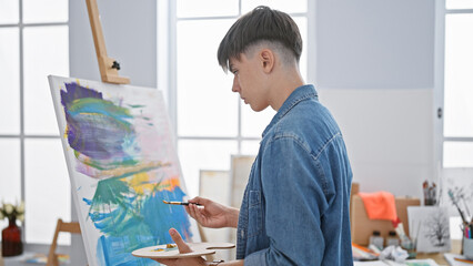 A young caucasian male artist wearing denim painting on a canvas in a bright studio room.