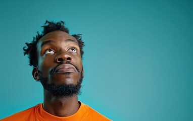 Fototapeta premium An African American man looks up with a puzzled expression, deep in thought, illuminated by a vibrant turquoise backdrop