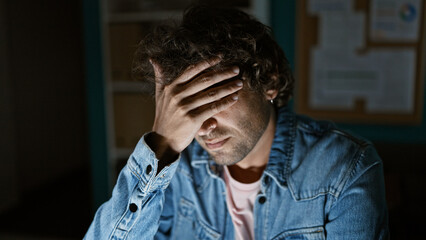 A young hispanic man in a denim jacket appears stressed in a dimly lit office setting.