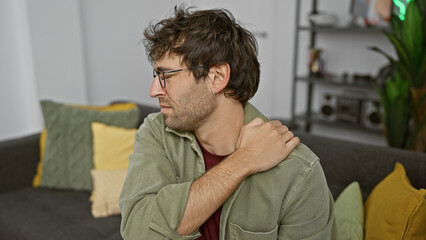 Fototapeta premium Handsome young man with beard and glasses touching his neck in pain in a modern apartment living room.