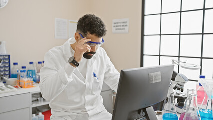 Bearded man analyzes data on computer in a modern laboratory setting with microscope and glassware present.